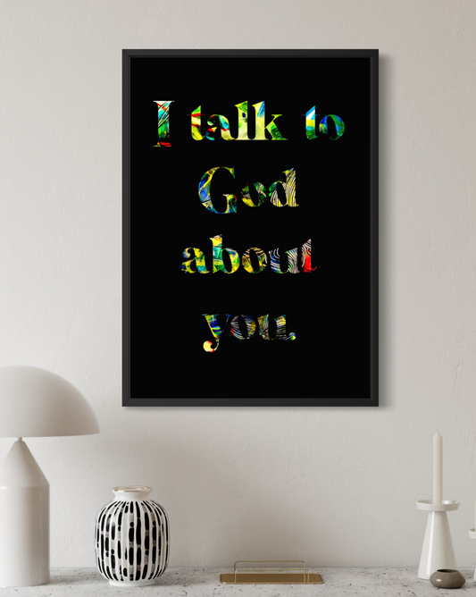 A framed wall art print with the text 'I talk to God about you' in multicolored letters on a black background, displayed in a modern room setting above a console table with a lamp, vase, and candle holder.