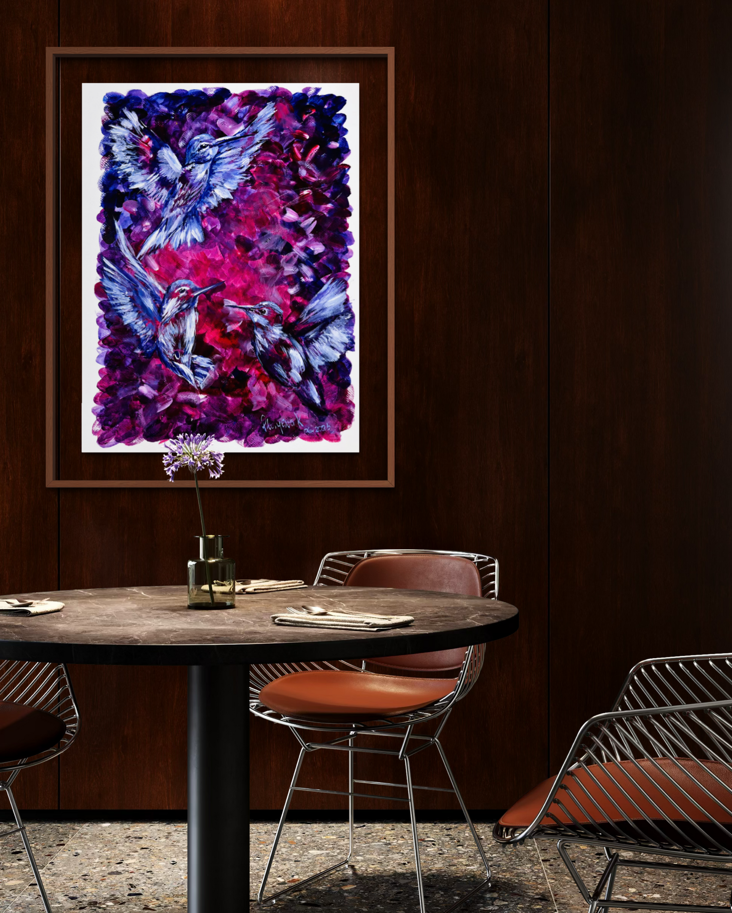 A framed abstract painting of hummingbirds in vibrant purple, pink, and blue tones, displayed on a dark wood-paneled wall above a round dining table with modern chairs.