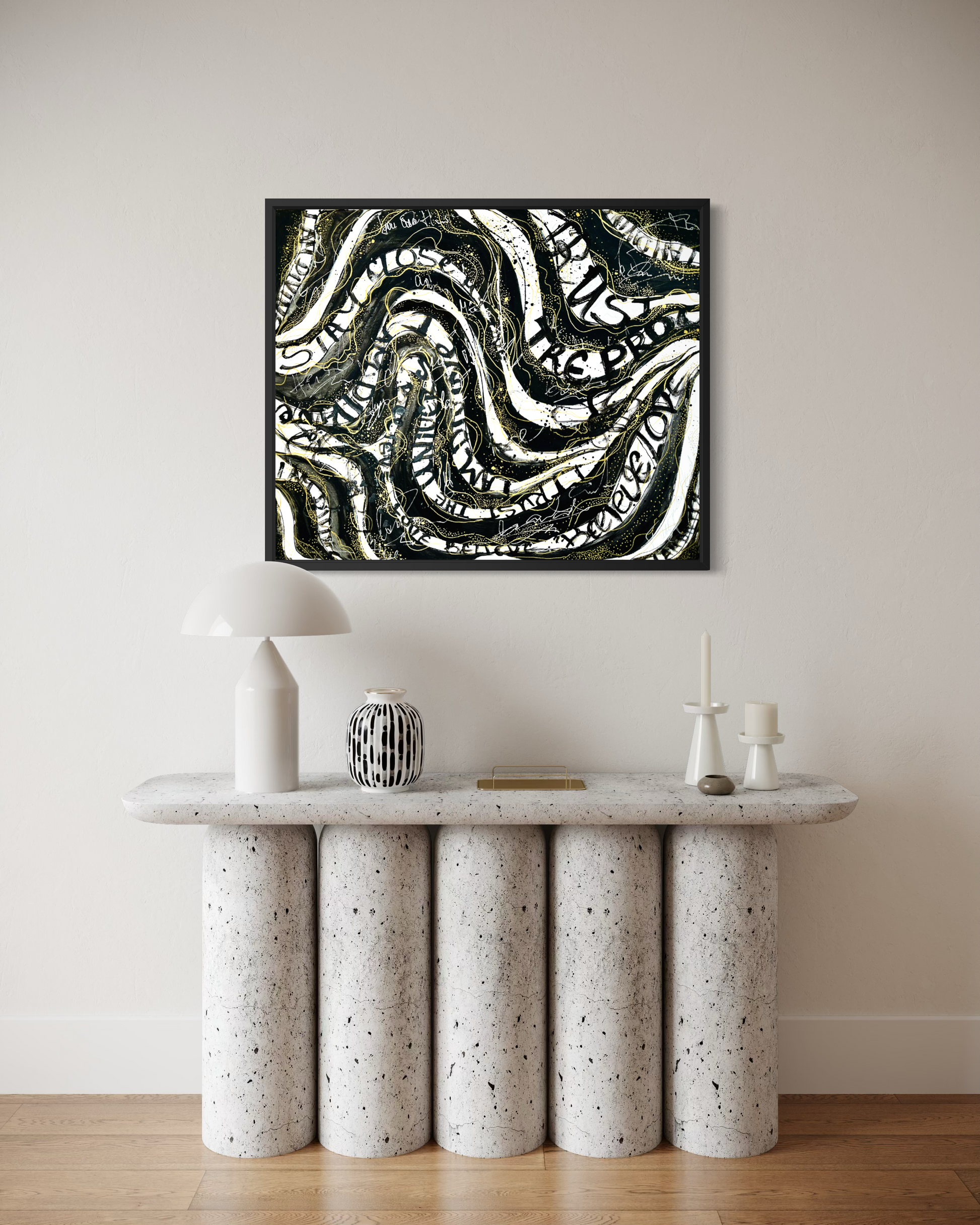 A framed abstract canvas print with black, white, and gray wavy lines hangs on a beige wall above a modern white and speckled stone console table with decorative objects.