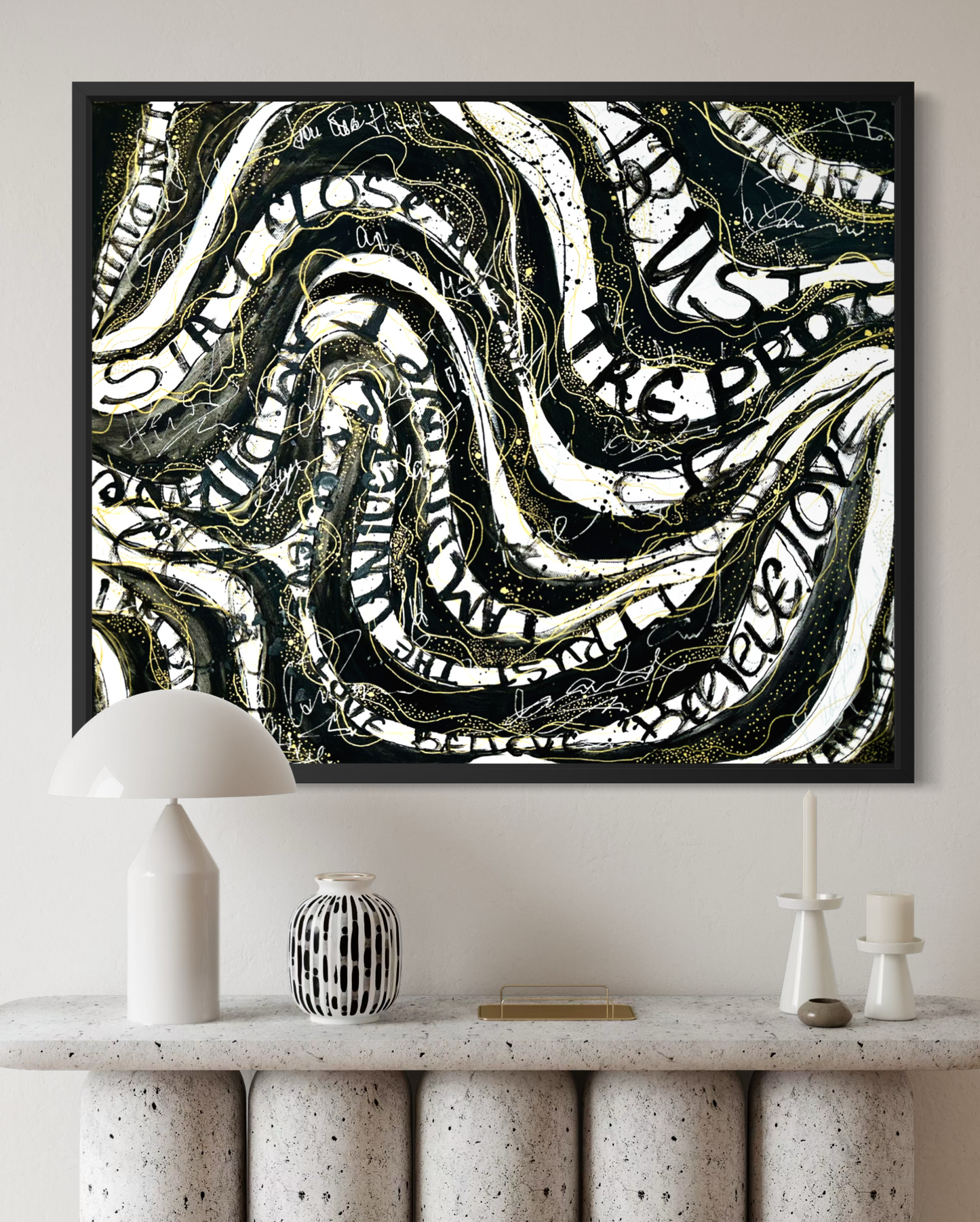 A framed abstract wall art print with swirling black, white, and gold text-like forms hangs above a console table in a styled interior. 