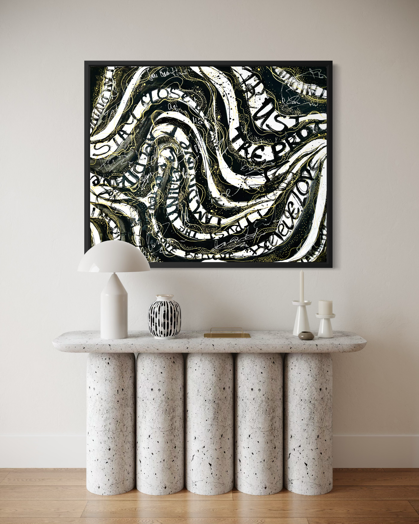 A framed abstract art print with black, white, and olive green wavy lines and text, displayed on a beige wall above a modern speckled stone console table with a white mushroom lamp and decorative objects.