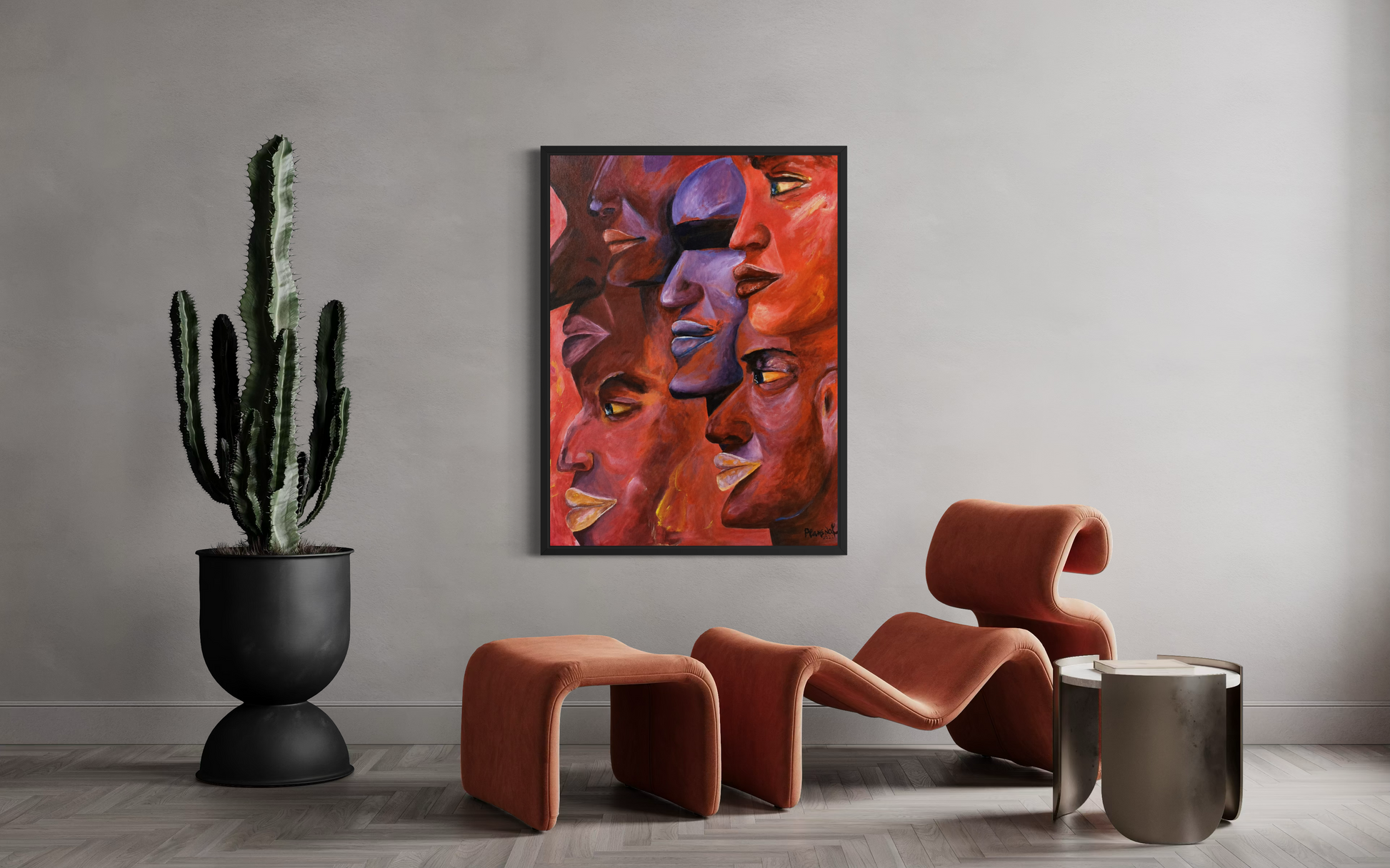 A framed abstract art print featuring multiple stylized faces in warm red, orange, and purple tones, displayed on a light gray wall in a modern living room setting with sculptural orange chairs, a potted cactus, and a metallic side table.