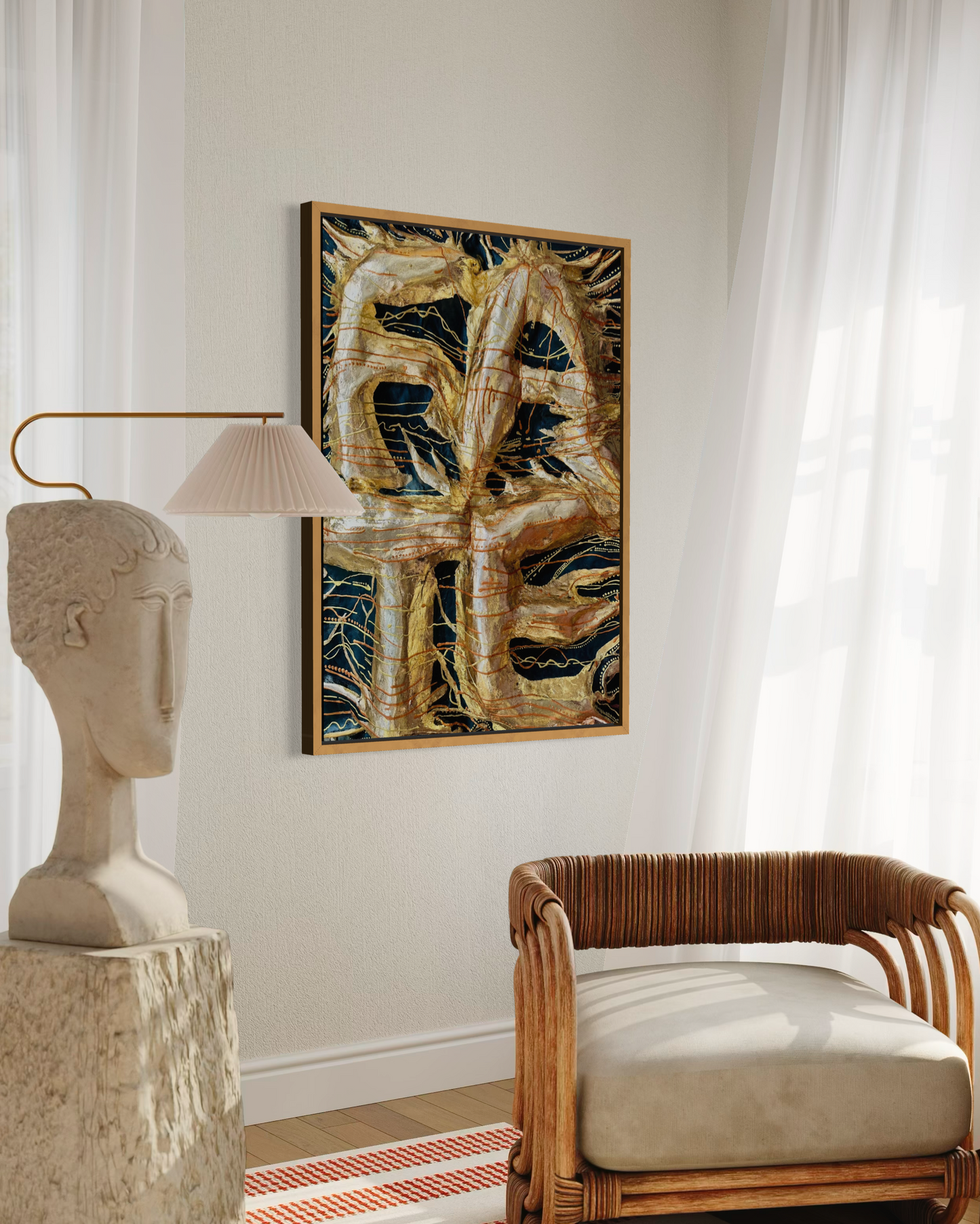 A framed abstract art print featuring gold, black, and beige organic shapes, displayed on a light beige wall in a styled room with a white lamp, a stone bust sculpture, and a rattan armchair.