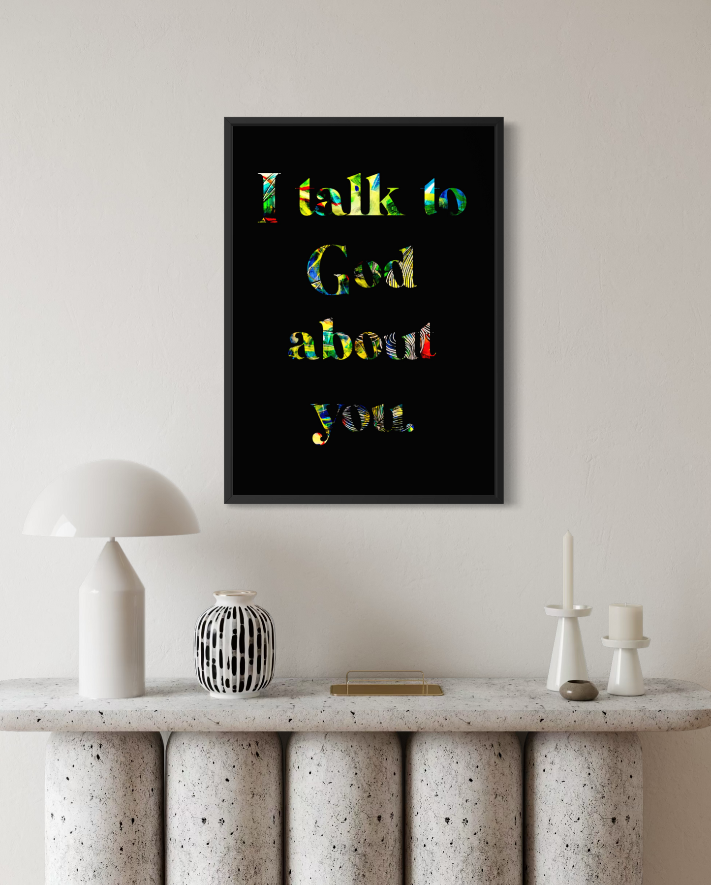 A framed wall art print with the text 'I talk to God about you.' in multicolored lettering on a black background, displayed above a console table with decorative objects in a styled interior setting.