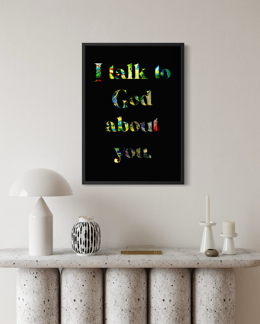A framed wall art print with the text 'I talk to God about you.' in multicolored lettering on a black background, displayed above a console table with decorative objects in a styled interior setting.