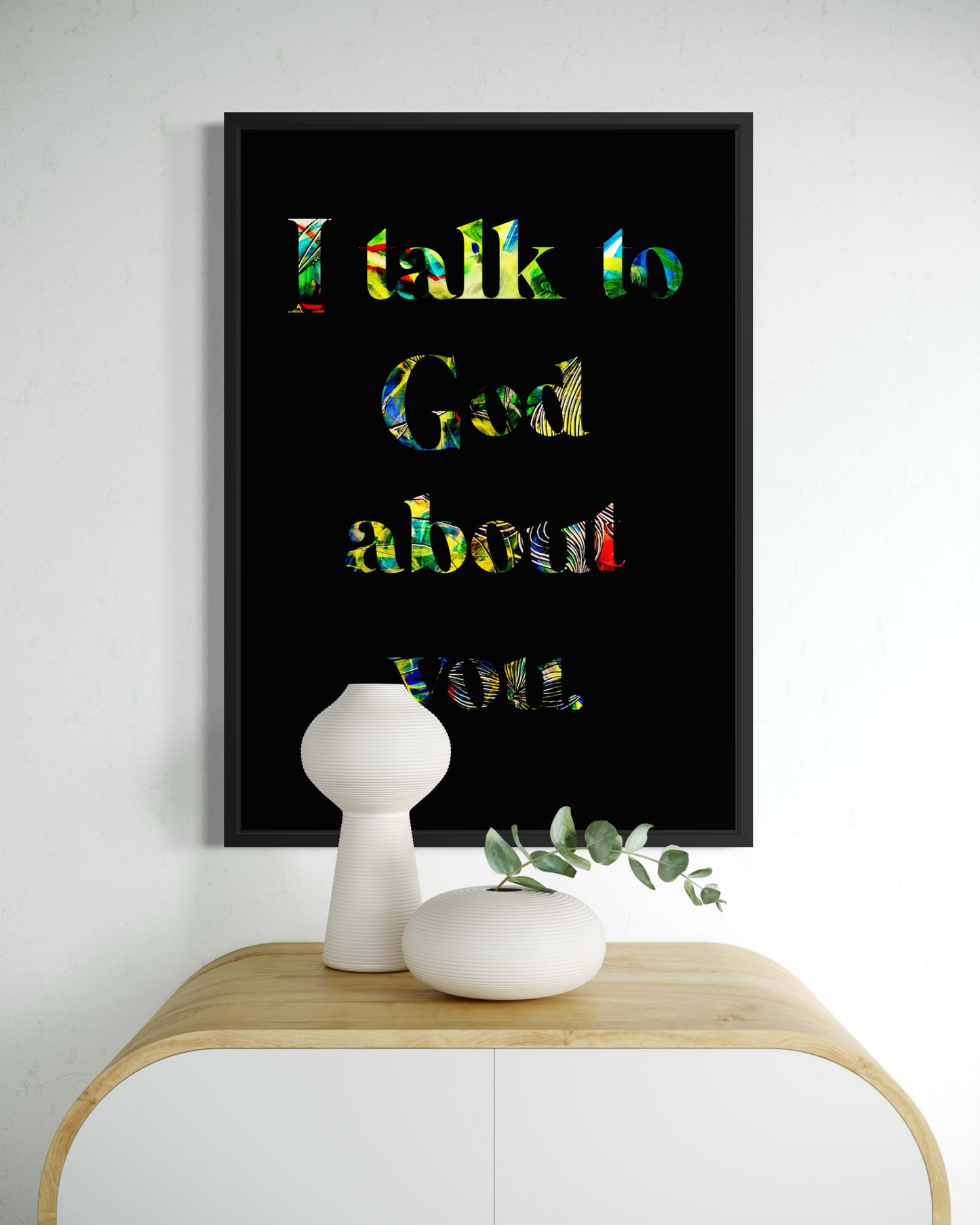 A framed wall art print with the text 'I talk to God about you' in multicolored lettering on a black background, displayed above a light wood console table with white decorative vases.