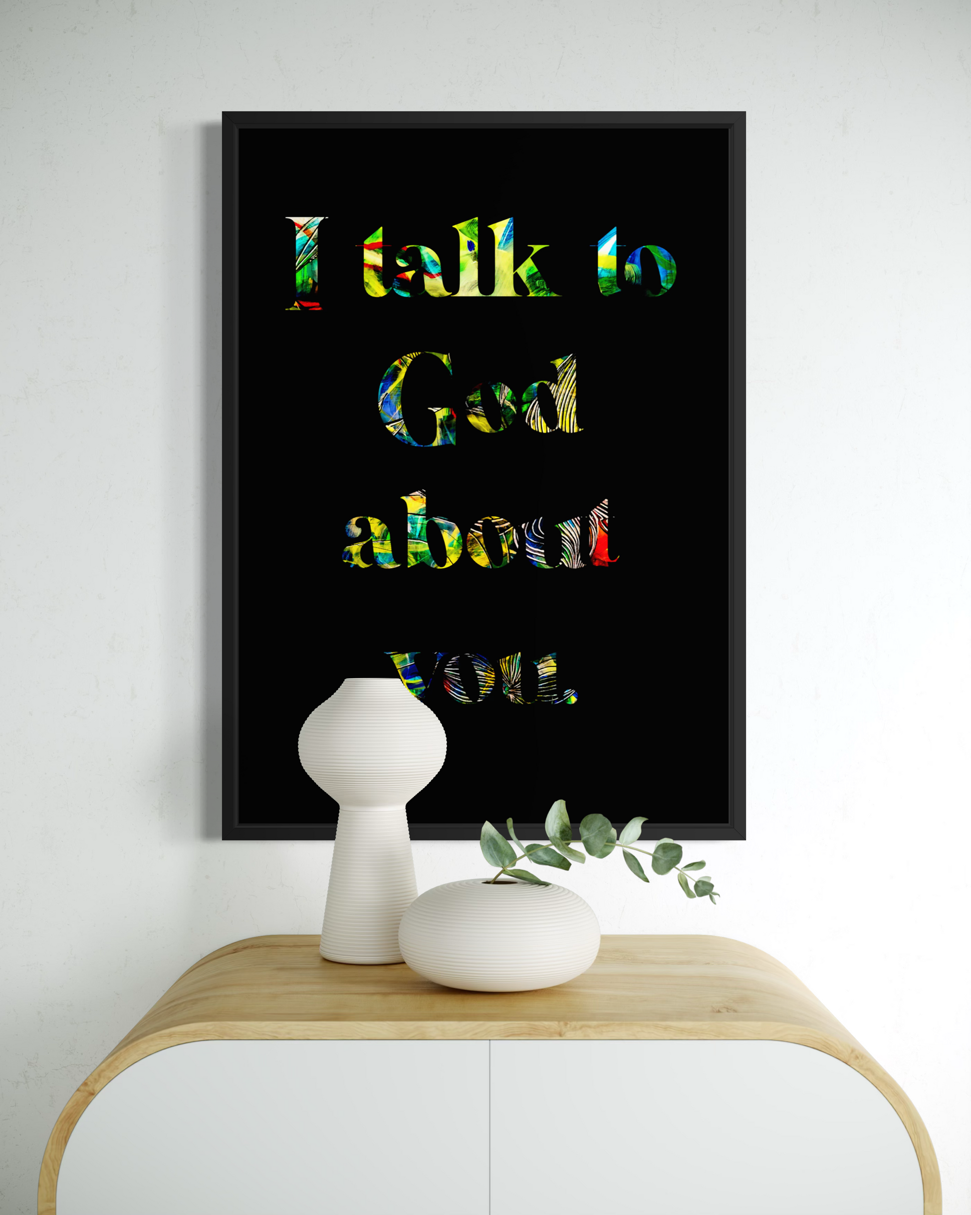 A framed wall art print with the text 'I talk to God about you' in multicolored lettering on a black background, displayed above a light wood console table with white decorative vases.