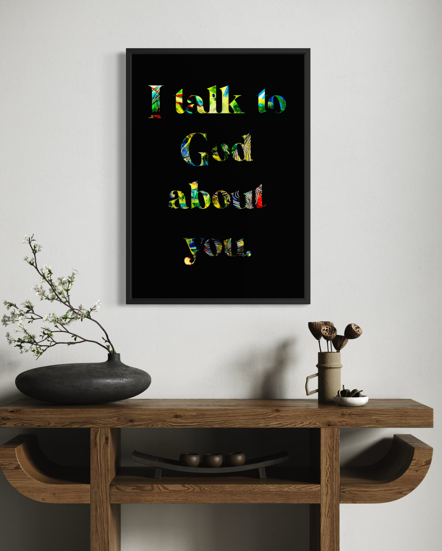 A framed wall art print with the phrase 'I talk to God about you.' in multicolored lettering on a black background, displayed above a wooden console table with decorative objects in a styled interior setting.