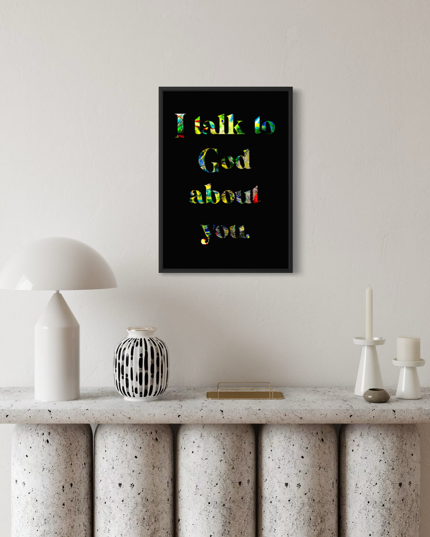 A framed wall art print with the text 'I talk to God about you.' in multicolored lettering on a black background, displayed above a stone console table with a white lamp and decorative objects in a styled interior setting.
