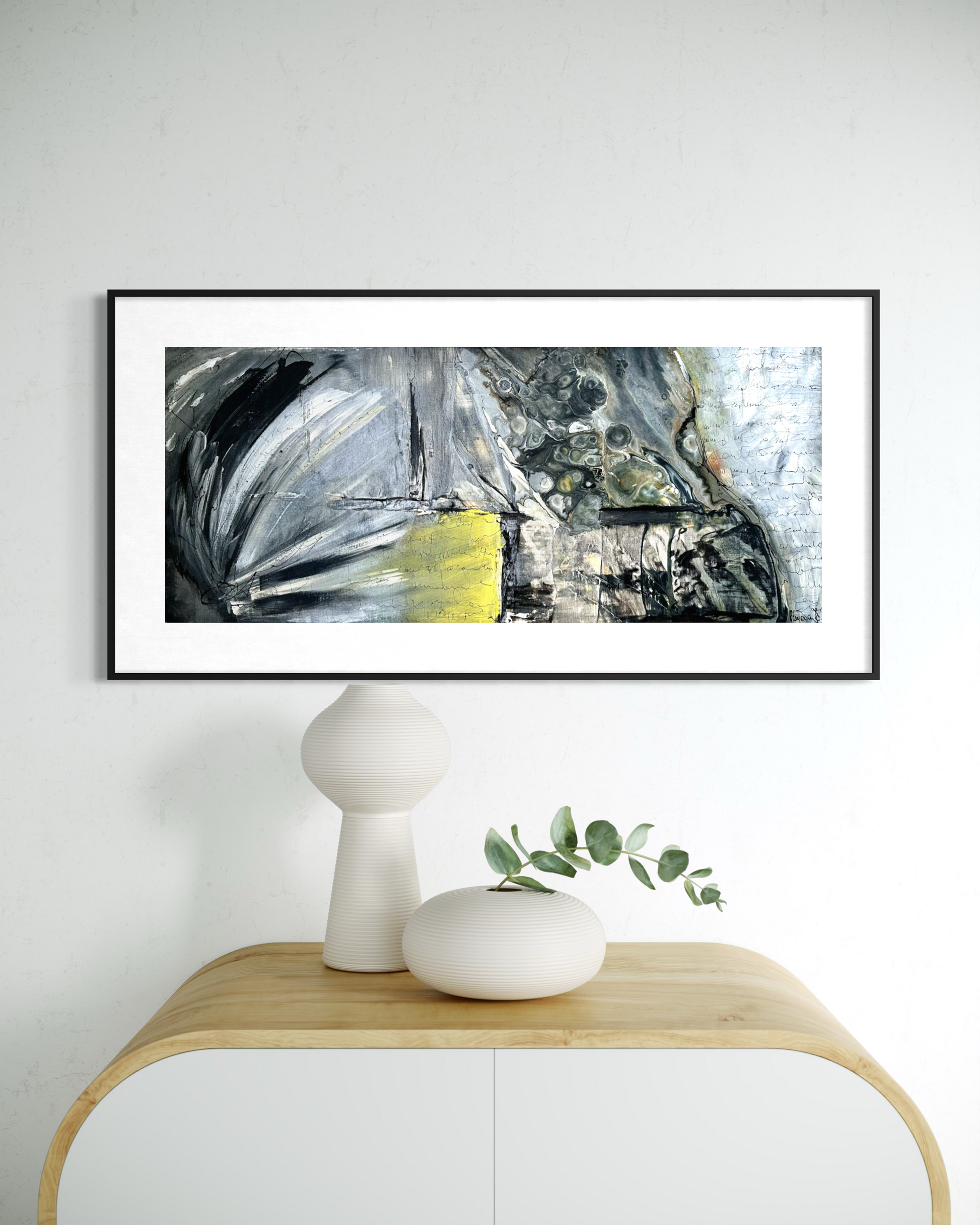A framed abstract canvas print with black, white, gray, and yellow brushstrokes, displayed on a white wall above a light wood console table with white ceramic vases.