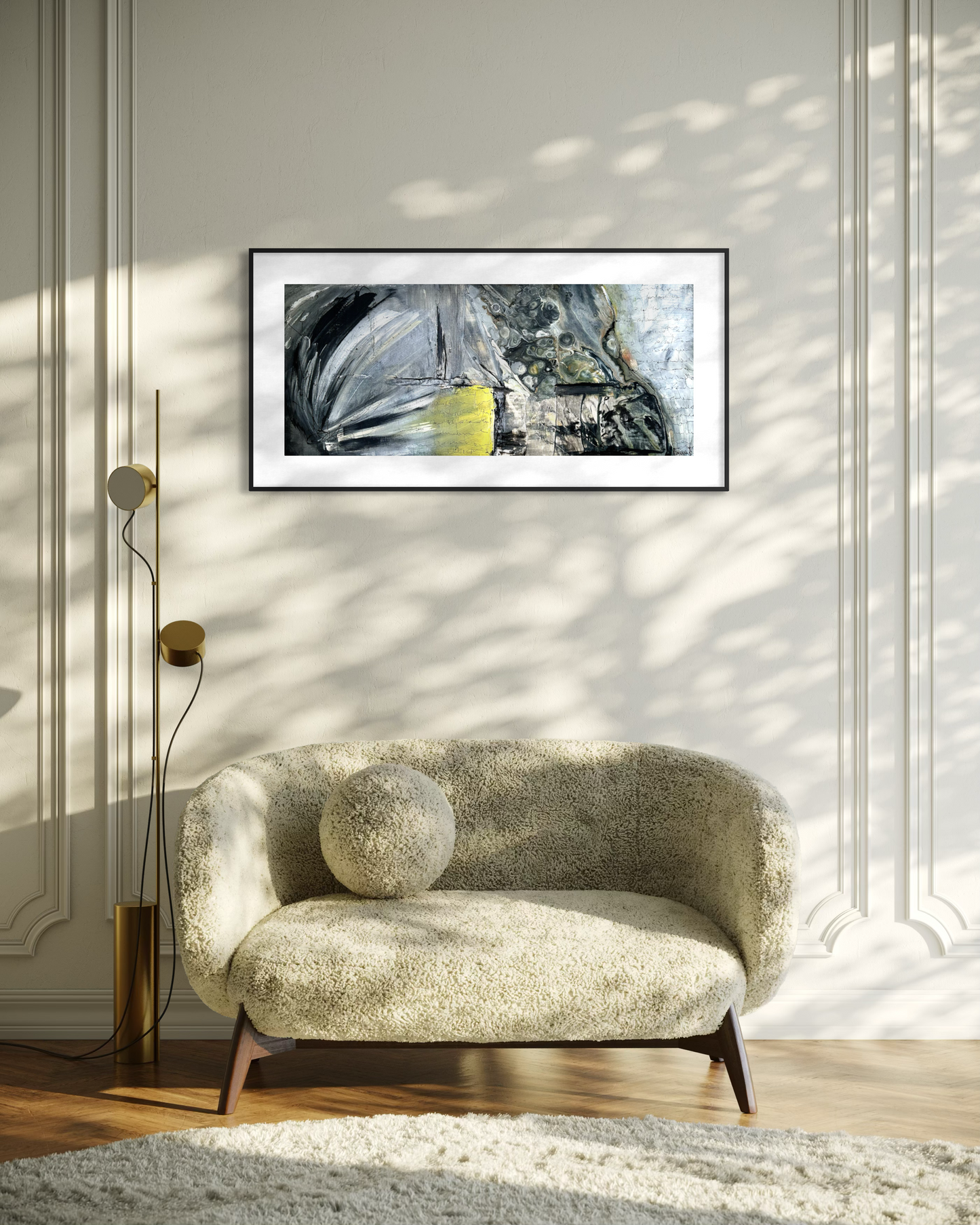 A framed abstract art print with black, gray, white, and yellow brushstrokes hangs on a cream paneled wall above a textured beige armchair in a sunlit room.