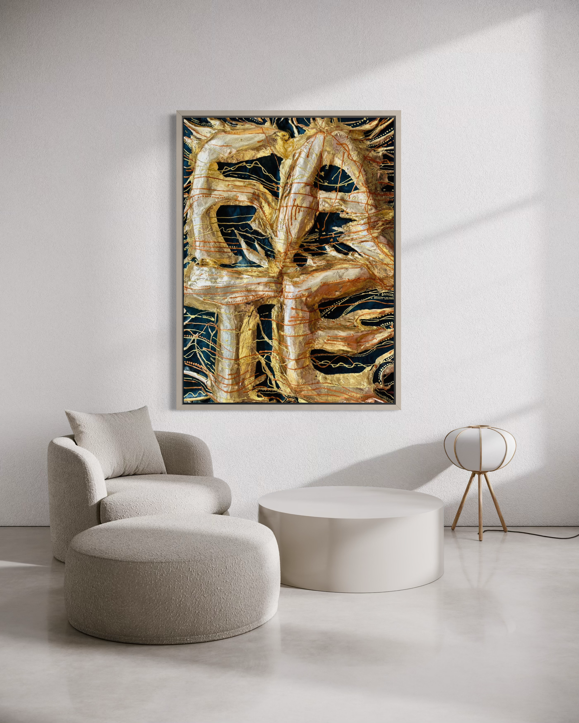 A framed abstract wall art print hangs on a white wall in a modern living room setting, featuring golden abstract shapes over a dark background. The room includes a light gray armchair, a round ottoman, a low white coffee table, and a small gold floor lamp.