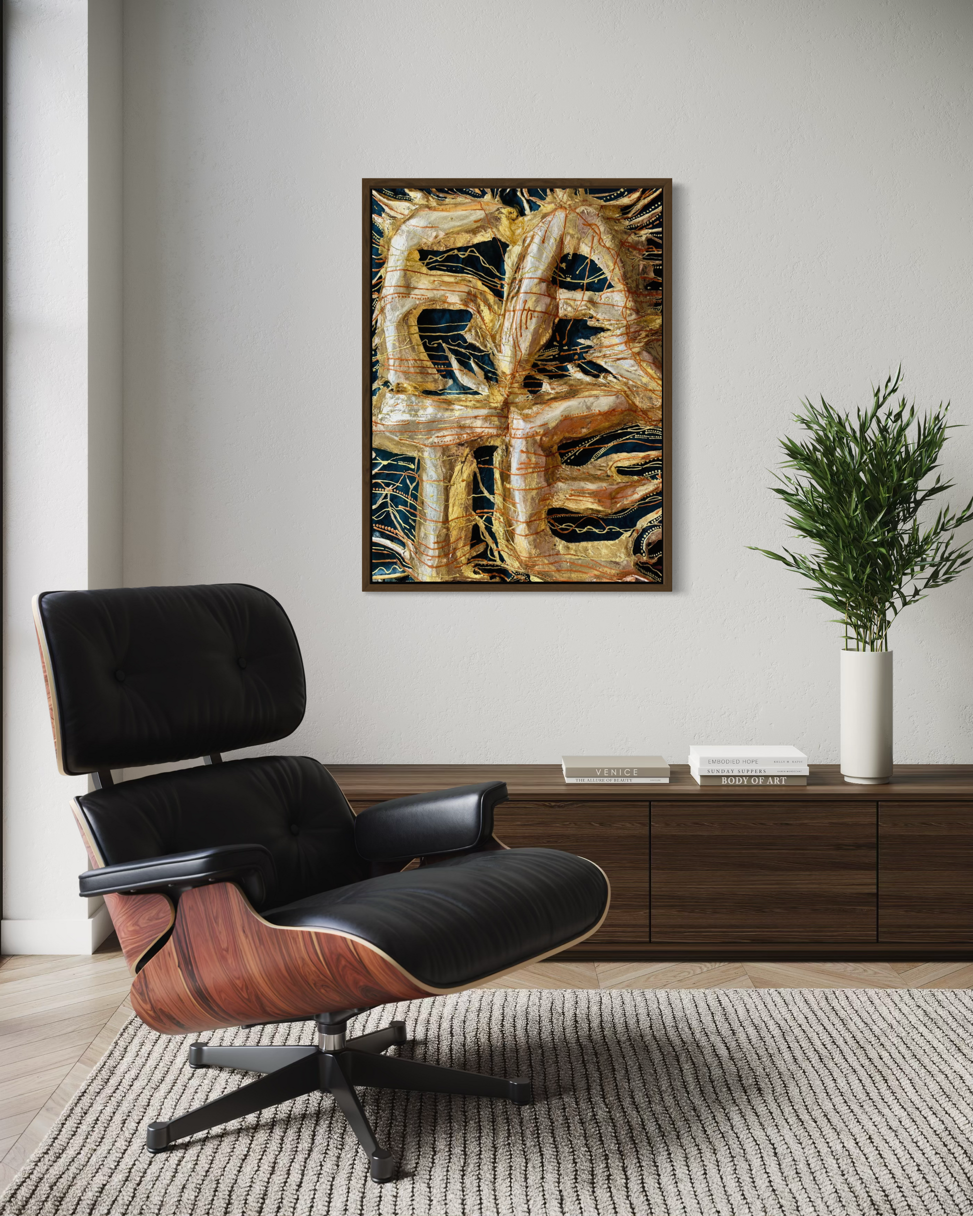 A framed abstract canvas print featuring textured gold, brown, and black brushstrokes, displayed on a light gray wall in a modern living room setting with a black leather and wood lounge chair, a wooden console table, and a potted green plant.